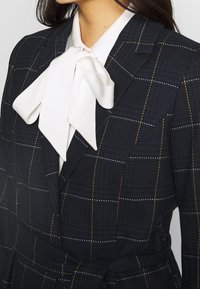 Granatowa marynarka w kratę z fakturą, z białą satynową muchą przy kołnierzu oraz ozdobnymi szwami w kolorze złotym i białym.