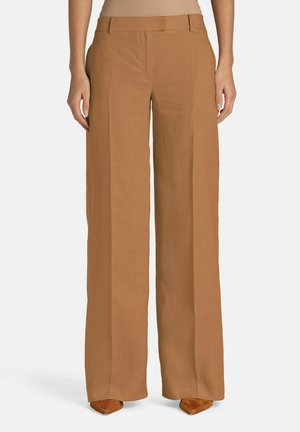 Kvinde iført camel-farvede bukser med høj talje og vide ben, kombineret med brune, spidse sko, stående foran en ensfarvet baggrund.