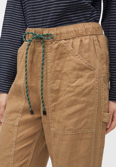 Pantalon décontracté marron avec grandes poches avant et cordon vert attaché en nœud, porté avec une chemise rayée bleu marine.