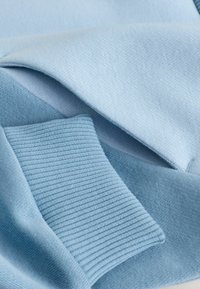 Hellblauer Pulloverstoff mit glatter Textur, der gerippte Bündchen und ein gefaltetes Design aufweist. Die Oberfläche ist ebenmäßig und leicht glänzend.