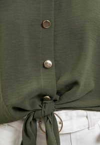 Blouse vert olive avec un lien à l'avant et quatre boutons dorés. Fabriquée en tissu léger avec une texture lisse et un léger éclat.