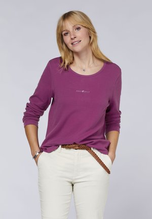 Purple long-sleeve shirt with a round neck and logo detail. Paired with light beige pants and a brown braided belt. Smooth cotton texture.