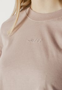 Šviesiai rožinė marškinėliai, pagaminti iš minkštos medvilnės. Yra sustiprinta apykaklė ir iškilęs "GUESS" logotipas krūtinėje. Sklandus, kasdienis dizainas.
