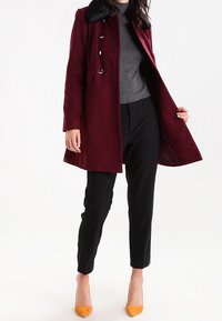 Manteau bordeaux avec col en fausse fourrure noire, design à double boutonnage, associé à un haut gris et un pantalon noir, avec des talons orange.