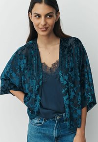 Flowing dark teal kimono with floral pattern and sheer fabric over a navy camisole featuring lace trim, paired with light blue denim jeans.