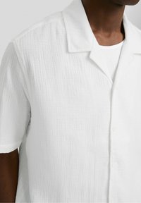 Uomo che indossa una camicia bianca a maniche corte con bottoni e tessuto strutturato, sopra a una maglia bianca a coste con collo rotondo, visibile dalle spalle a metà busto.