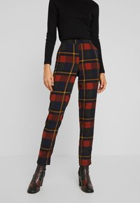 Kvinna iklädd högmidjade, smala rutiga byxor i röd, marinblå och olivgrön med senapsfärgade linjer, i kombination med en svart långärmad topp och svarta ankelstövlar.