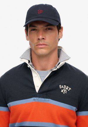 Man wearing navy blue and orange striped rugby shirt with popped collar and navy blue cap with red logo on a white background.