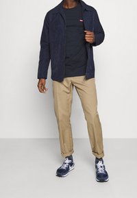 Marinblå zip-up jacka med texturerad yta, marinblå t-shirt, khakifärgade byxor och marinblå sneakers med vita accenter och logotyp.