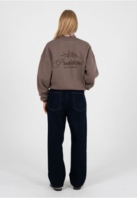 Frau mit langen blonden Haaren, trägt ein locker sitzendes braunes Sweatshirt mit dem Aufdruck "Prohibited 10119 Berlin" und dunkle Jeans mit weitem Bein, steht mit dem Rücken zur Kamera.
