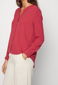 Camicia rossa a maniche lunghe con scollatura a V, chiusura con bottoni, vestibilità rilassata e tessuto morbido. Abbinata a pantaloni chiari, comodi e larghi.