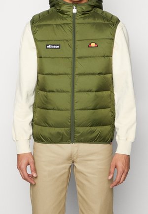 Zielona pikowana kamizelka z kapturem, zapinana na przedni zamek, z czarną naszywką z logo oraz małym czerwono-białym akcentem logo. Noszona na kremowej koszuli z długim rękawem.