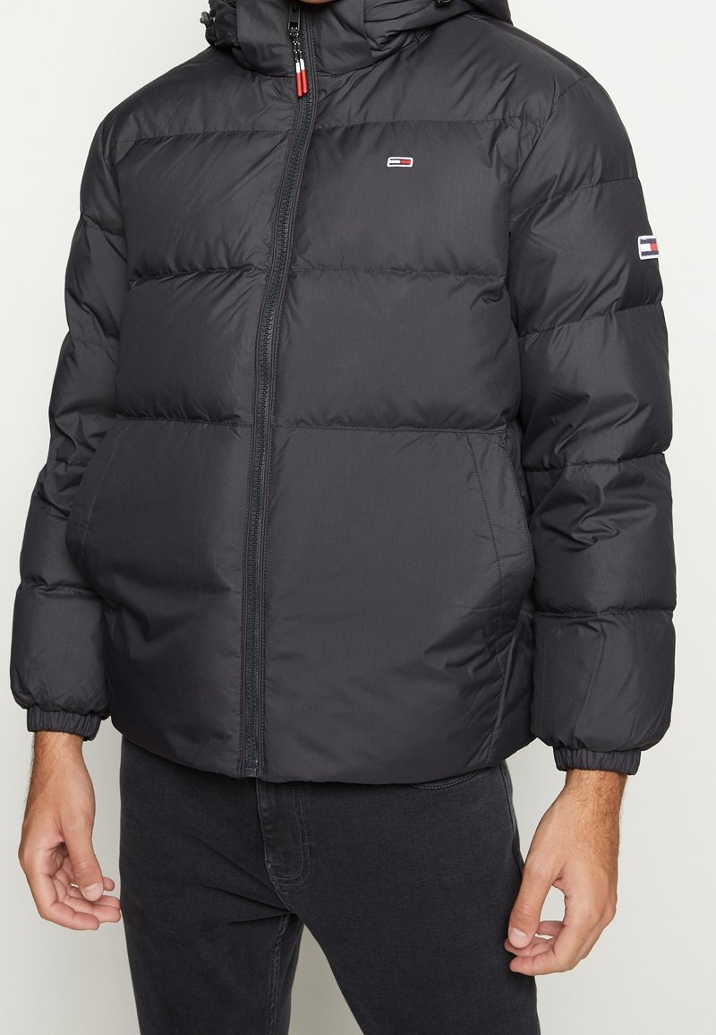 Veste matelassée noire avec capuche portée sur un pantalon foncé, avec de petits logos de marque rectangulaires rouges, blancs et bleus sur la poitrine et la manche.