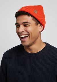 Orange knit beanie with a ribbed texture and a small leather patch. Worn with a dark navy knitted sweater. Smiling individual.