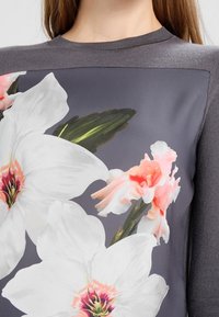 Grå långärmad topp med ett blommönster som har stora vita blommor och rosa accenter på en mörkgrå bakgrund, med en strukturerad stickad krage.