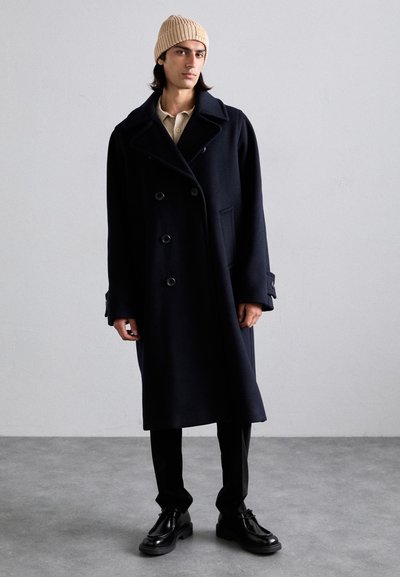 Manteau en laine bleu marine à double boutonnage, avec un grand col cranté et des accents de boutons, porté sur un pantalon noir et des chaussures brillantes, associé à un bonnet en tricot beige.