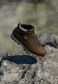 Bota de senderismo de cuero marrón con acentos grises, con ojales de metal, cordones y una suela de goma texturizada, situada contra un fondo rocoso.