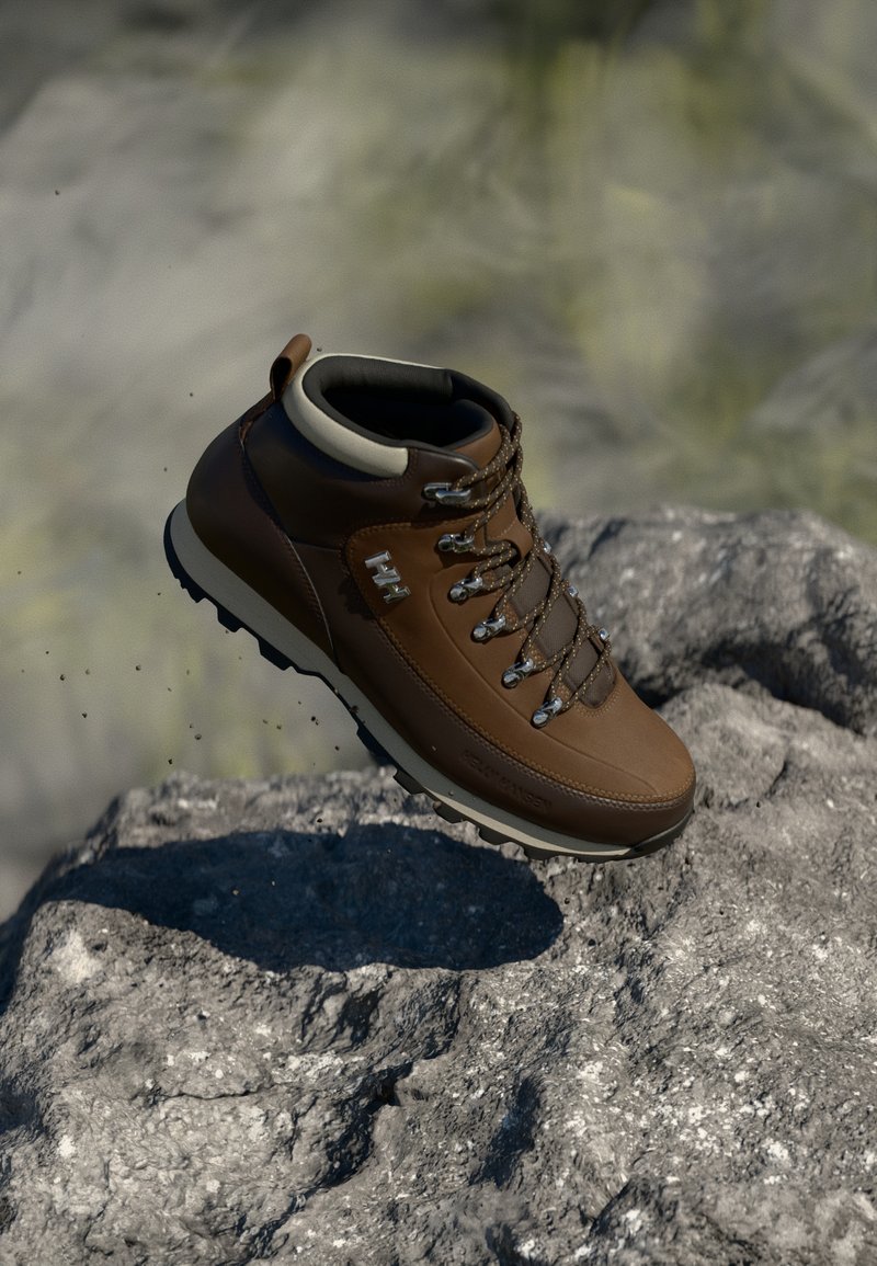 Bota de senderismo de cuero marrón con acentos grises, con ojales de metal, cordones y una suela de goma texturizada, situada contra un fondo rocoso.