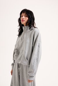 Γκρι κοντό φούτερ με μισό φερμουάρ, μπροστινή τσέπη και γυαλιστερά γράμματα κατά μήκος του μανικιού. Φτιαγμένο από μαλακό ύφασμα με άνετη εφαρμογή.