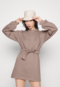 Robe taupe à coupe décontractée, manches longues et ceinture nouée à la taille. Associée à un chapeau bob beige clair. Texture lisse.