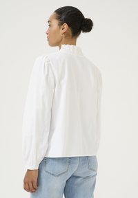 Blouse blanche à manches longues avec un col à volants, des manches bouffantes et une coupe décontractée. Tissu en coton lisse, associée à un jean bleu clair.