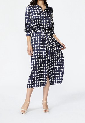 Vrouw draagt een midi-jurk met knopen in marineblauw en wit patroon, lange mouwen, getailleerde taille en witte sandalen met hak, staand op een witte achtergrond.