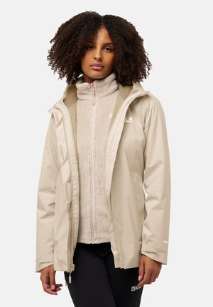 Vrouw met krullend haar die een beige waterdichte jas draagt over een lichtbeige fleece jas en zwarte broek, staand tegen een effen achtergrond.