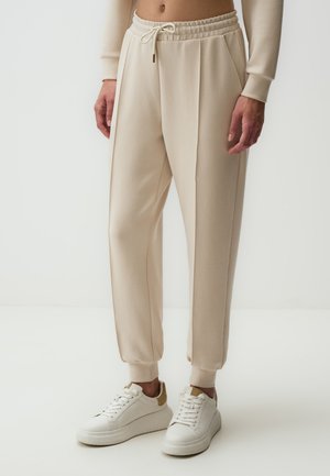 Pantaloni sportivi beige con vita regolabile tramite stringa, design plissettato e caviglie elastiche, abbinati a sneakers bianche con un accento color beige.