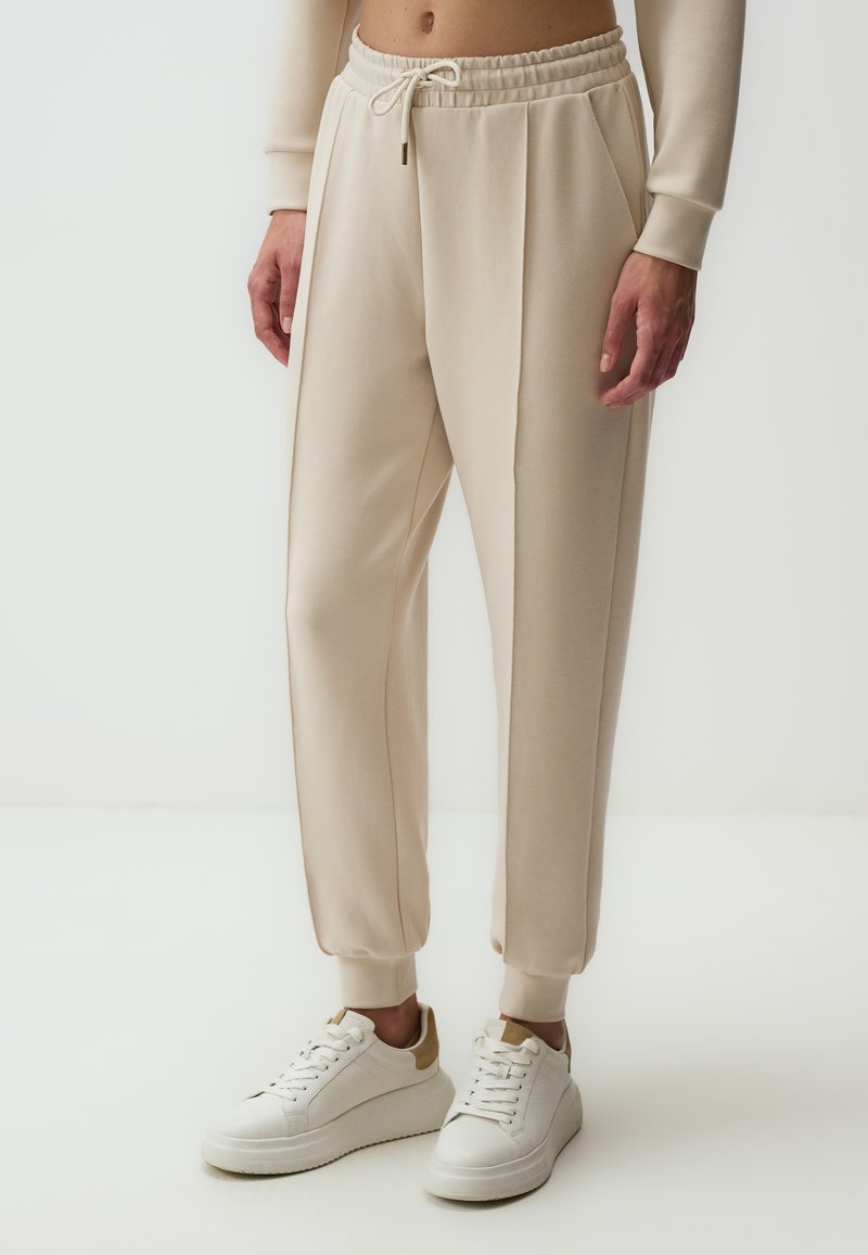 Beige sweatpants met een koord in de taille, geplooid ontwerp en boordjes aan de enkels, gecombineerd met witte sneakers met een tan accent.
