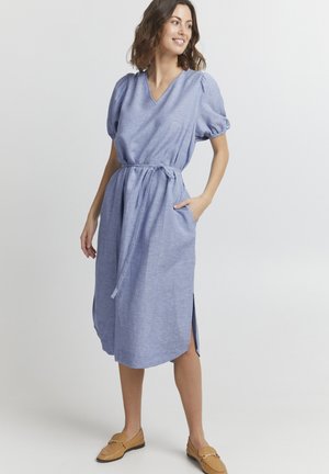 Hellblaues Leinenmischkleid mit Puffärmeln, V-Ausschnitt, tailliertem Gürtel, seitlichen Taschen und seitlichen Schlitzen. Kombiniert mit beigen Slippern.