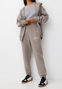 Ljusgrå hoodie och matchande sweatpants, med en logotyp. Svartvita sneakers med gummisula fulländar den avslappnade outfiten.