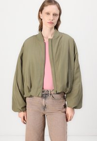 Olivově zelená bomber bunda, uvolněný střih, zapínání na zip, oversized rukávy. Oblékaná přes růžový top a spárovaná se světle barevnými džínami.