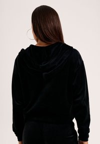 Schwarze Samt-Hoodie mit lockerem Schnitt, hinten mit einer hängenden Kapuze und langen Ärmeln, von hinten betrachtet. Glatte Textur.