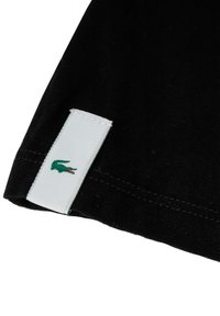 Orlo di una camicia di tessuto nero con un'etichetta bianca che presenta un logo di coccodrillo verde. Il tessuto ha una superficie liscia con dettagli di cucitura precisi.