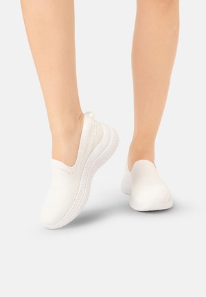 Een paar blote benen met witte slip-on gebreide sneakers met dikke gestructureerde zolen op een lichtgrijze achtergrond.