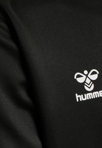 Svart idrottströja tillverkad av slätt, lättviktigt tyg, med en vit logo som har en stiliserad bi och varumärkesnamnet "hummel" tydligt visas.