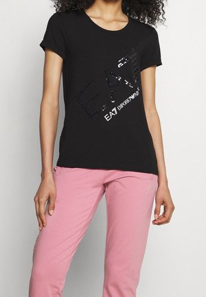 Femme portant un t-shirt noir à manches courtes avec le logo brillant "EA7 Emporio Armani" et un pantalon rose clair sur un fond uni.