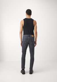 Černý bezrukávový top a tmavě šedé slim-fit džíny s koženým zadním výložníkem, zobrazené zezadu na jednobarevném bílém pozadí.