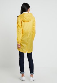 Geel waterdicht regenjack met capuchon, elastische manchetten en een geaccentueerde taille; gedragen over donkere spijkerbroeken en witte sneakers.