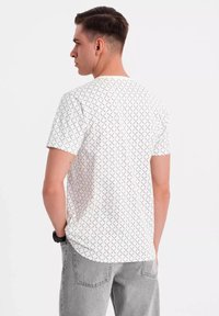 Bílá tričko s krátkým rukávem, které má opakující se černý diamantový vzor. Bavlněný materiál, kulatý výstřih, uvolněný střih s zakřiveným lemem.