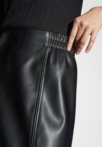 Jupe en cuir noir avec une texture lisse, présentant une taille froncée et des détails de coutures sur le côté, avec une main partiellement visible reposant sur le tissu.