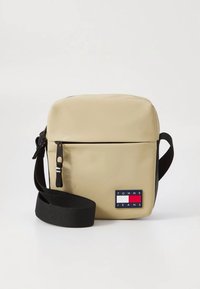 Beige axelremsväska med en mjuk, matt yta, utrustad med en svart justerbar rem och en färgglad logotypmärkning på framsidan.