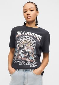 Νεαρό άτομο με πλεξούδες, φοράει μαύρο t-shirt Billabong με σχέδιο λουλουδιών και βουνών και ανοιχτόχρωμα τζιν, με τα χέρια στις τσέπες.