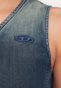 Farmer mellény fakó kék színben, mély V-nyakkivágással. Kék hímzett logó a mellkasnál és látható varrások az éleken.