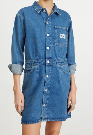 Robe en jean bleu à boutons avec manches retroussées, poche poitrine avec écusson logo et détail à la taille devant, portée par une personne.