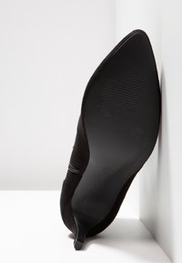 Chaussure à talon kitten en daim noir à bout pointu, positionnée sur le côté, montrant la semelle en caoutchouc texturée et un détail de fermeture éclair sur le dessus.
