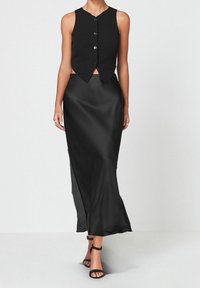 Schwarzes, geripptes Top mit fünf Knöpfen, kombiniert mit einem fließenden schwarzen Maxirock aus Satin, der einen seitlichen Schlitz hat, und dünnen schwarzen Riemchen-Heels.