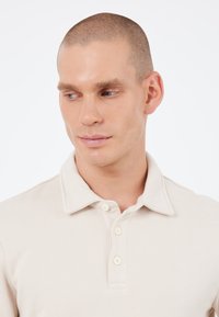 Polo a maniche corte di colore beige chiaro con colletto, tre bottoni e una texture liscia. Presenta un design aderente.