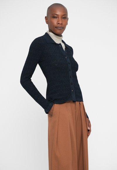 Gilet en laine côtelée marine, avec fermeture à boutons, coupe ajustée. Porté par-dessus un col roulé rayé. Pantalons beiges plissés. Texture lisse, col classique.