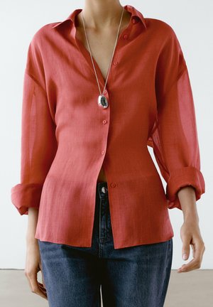 Persona che indossa una camicia rossa ampia con bottoni e maniche arrotolate, jeans scuri e una collana con ciondolo d'argento su uno sfondo semplice.